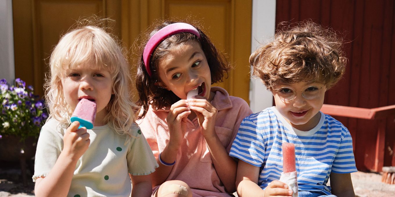 Tre barn sitter utomhus i solen på sommaren och äter glass.