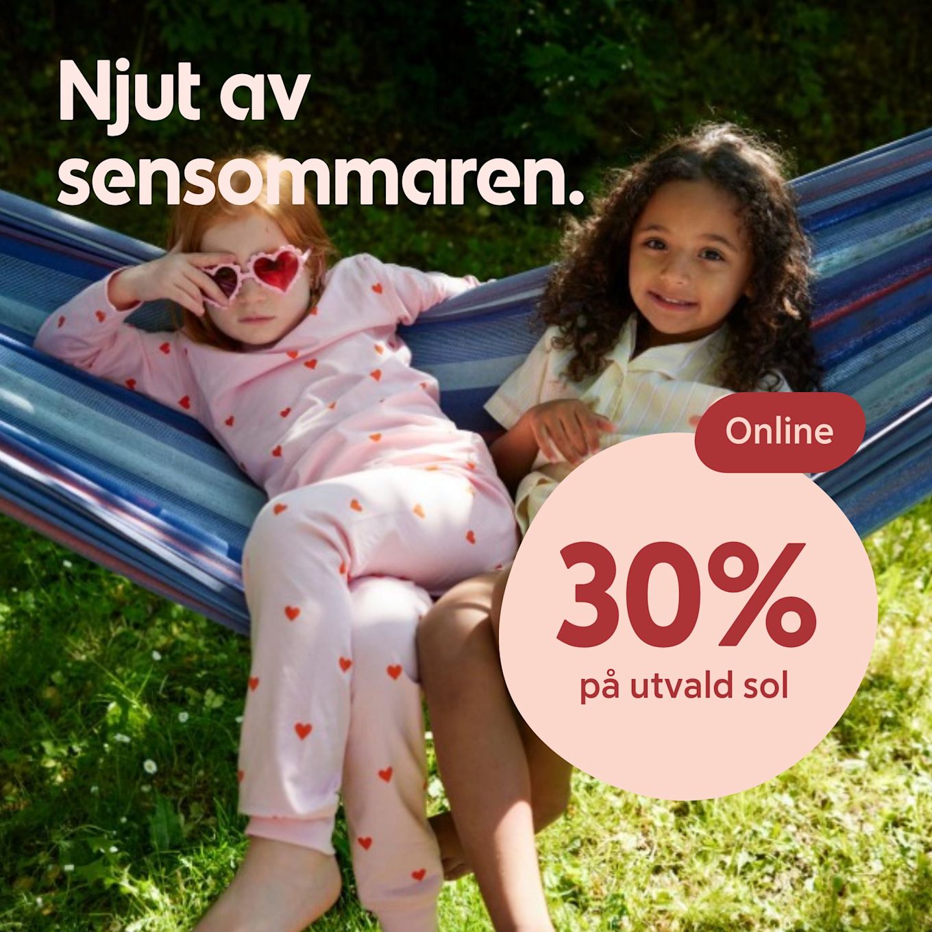 Två barn ligger avslappnat i en hängmatta utomhus på gräs, ett barn bär pyjamas med hjärtan och solglasögon. 30% på utvald sol online.