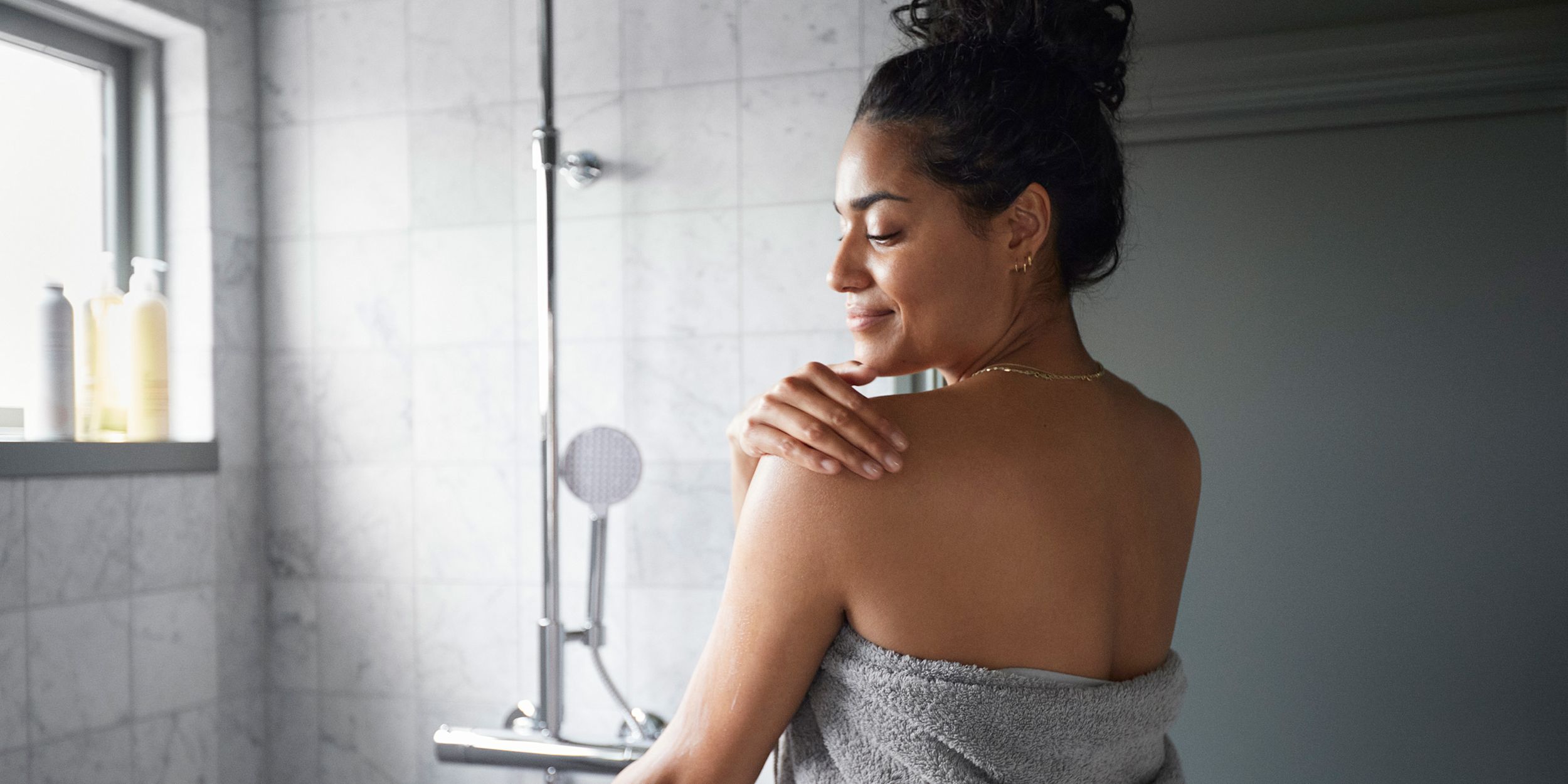 Kvinna står utanför duschen med handduk och smörjer in sin axel.