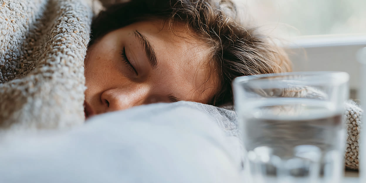 Sjuk tonåring, sovandes och nedbäddad i sängen med täcket långt uppdraget. Ett glas vatten syns på sängbordet. 