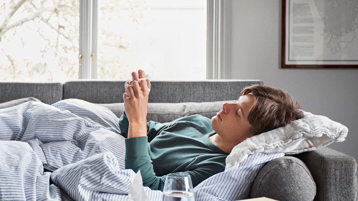 Förkyld ung man ligger i soffan med täcke och kuddar och tittar på sin mobiltelefon.