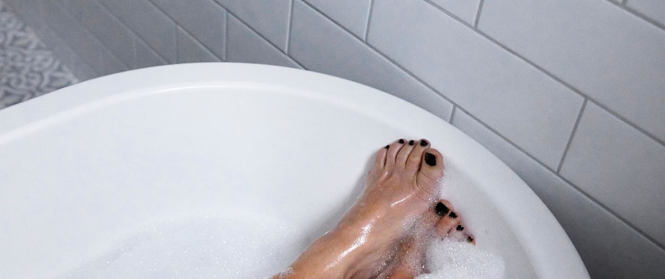 Två fötter syns sticka fram i ett badkar fullt med bubblor, fötterna har målade tånaglar och bilden ger uttryck för self care