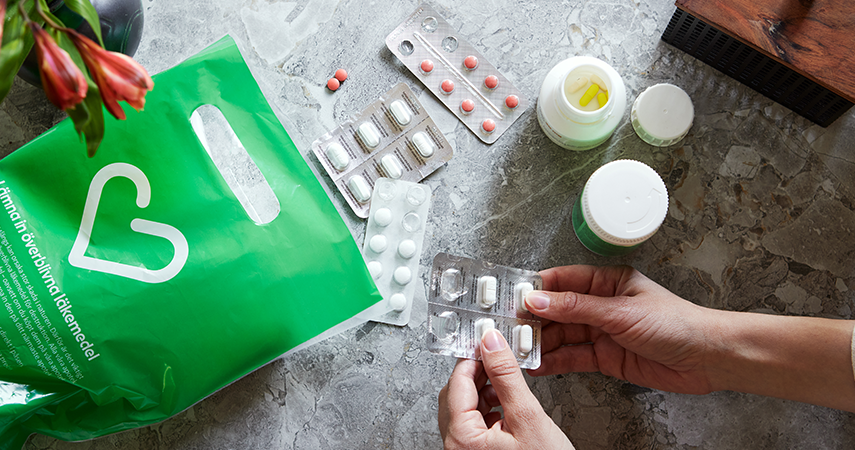 På ett grått marmorbord ligger röda, gula och vita tabletter och kapslar i burk och blister. Ett handpar håller i en tablettkarta med vita tabletter. En grön Apotek Hjärtat påse med texten: Lämna in överblivna läkemedel, är placerad bredvid är placerad bredvid.