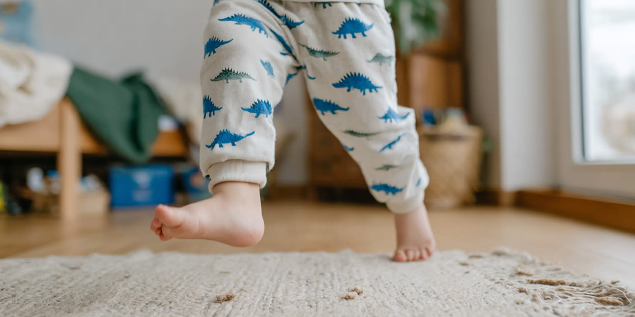Barn springer över en ljusbeige matta, barfota i sitt rum. Barnet har en ljust färgad pyjamas med gröna och blå dinosaurier på. 