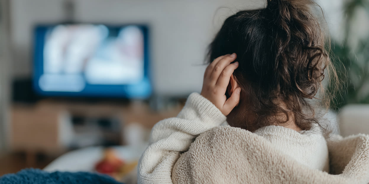 Liten flicka med mörkt lockigt hår uppsatt i toffs sitter i mjukisdress framför tv:n i soffan. Hon håller sig för ena örat.