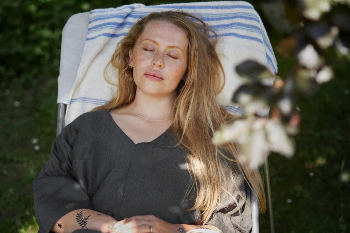 Kvinna med långt ljust hår sitter lutad mot en blå och vitrandig handduk i en solstol i skuggan. Hon är klädd i en mörkgrå, tunn och luftig klänning. Hon befinner sig i skuggan och vilar med slutna ögon. 