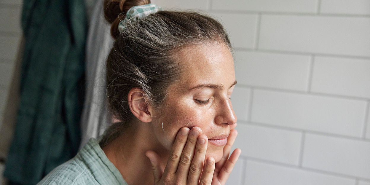 Kvinna i badrumsmiljö som smörjer in sina kinder med ansiktskräm.