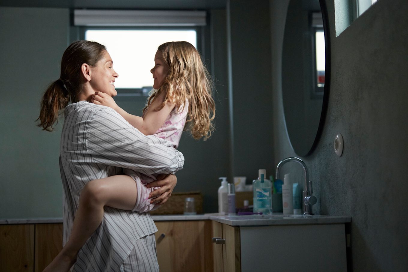 Mamma och dotter är i badrummet. Mamma håller sin dotter i famnen och båda skrattar.