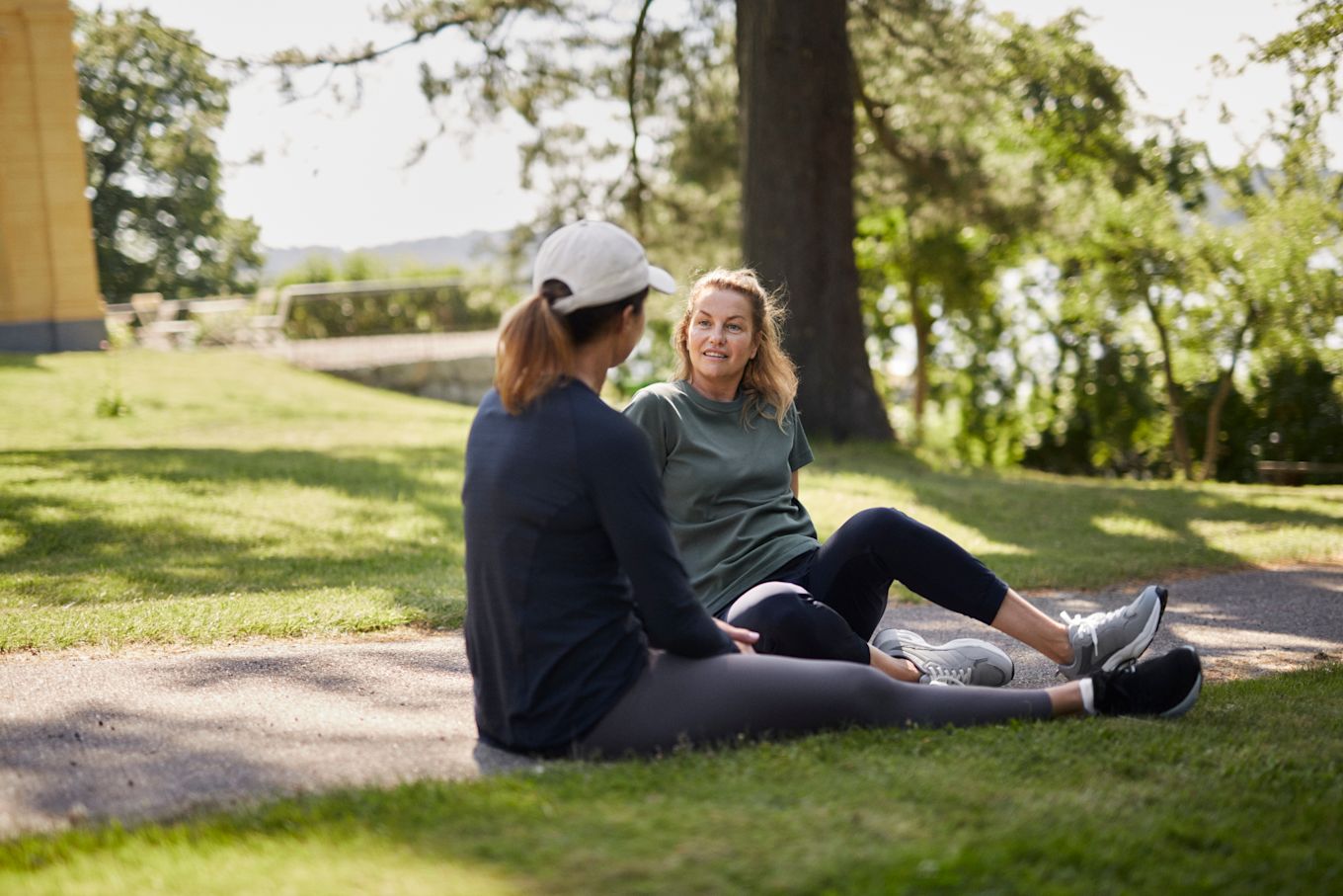 Två kvinnor sitter utomhus i gräset och pratar med varandra, iklädda träningskläder.