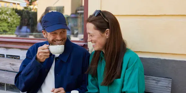 Man och kvinna sitter ute mot en vägg och fikar. De ler och verkar ha trevligt. Mannen har keps och dricker varm dryck. Kvinnan har solglasögon på huvudet. 