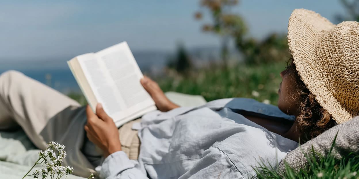 Kvinna med solhatt ligger i gräset och läser en bok.