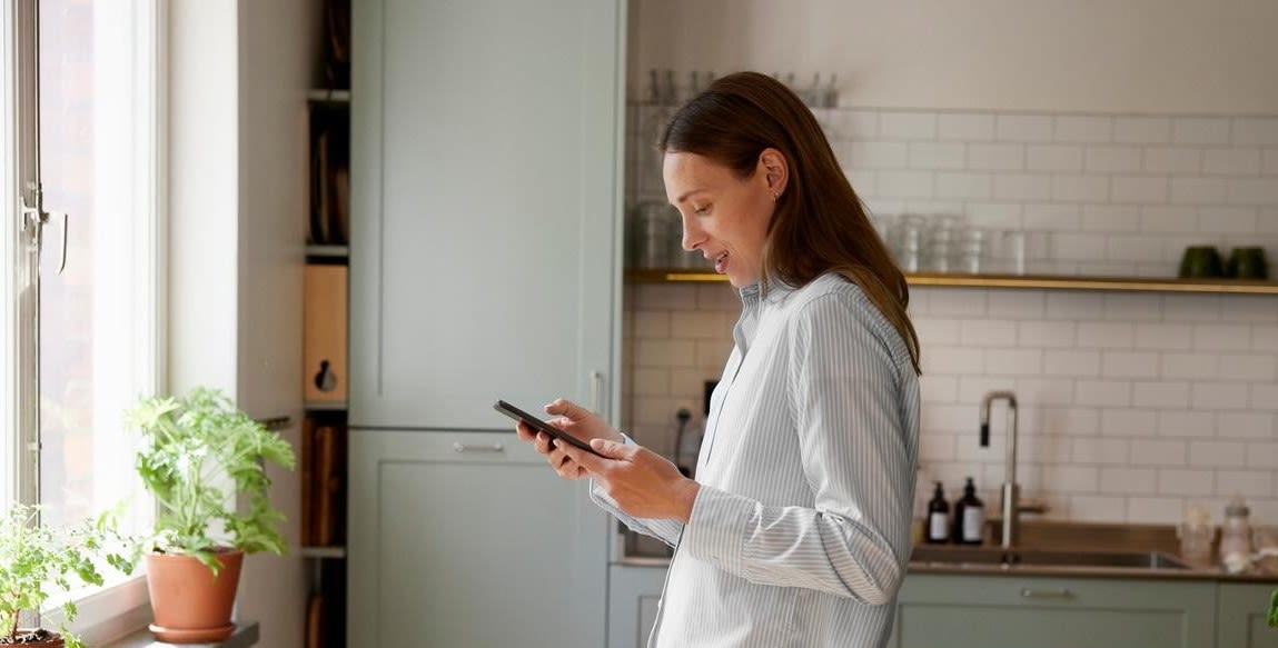 Kvinna med långt mörk utsläppt hår står i ljus köksmiljö framför ett fönster och tittar ned i sin mobil. hon har en tunn och ljus långärmad skjorta på sig.