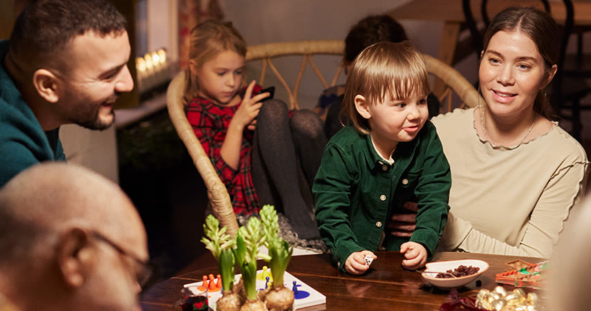 En familj med barn och vuxna sitter samlade runt matbordet och pratar, rummet är pyntat för jul med hyacinter och ljusstakar. På bordet står julgodis.