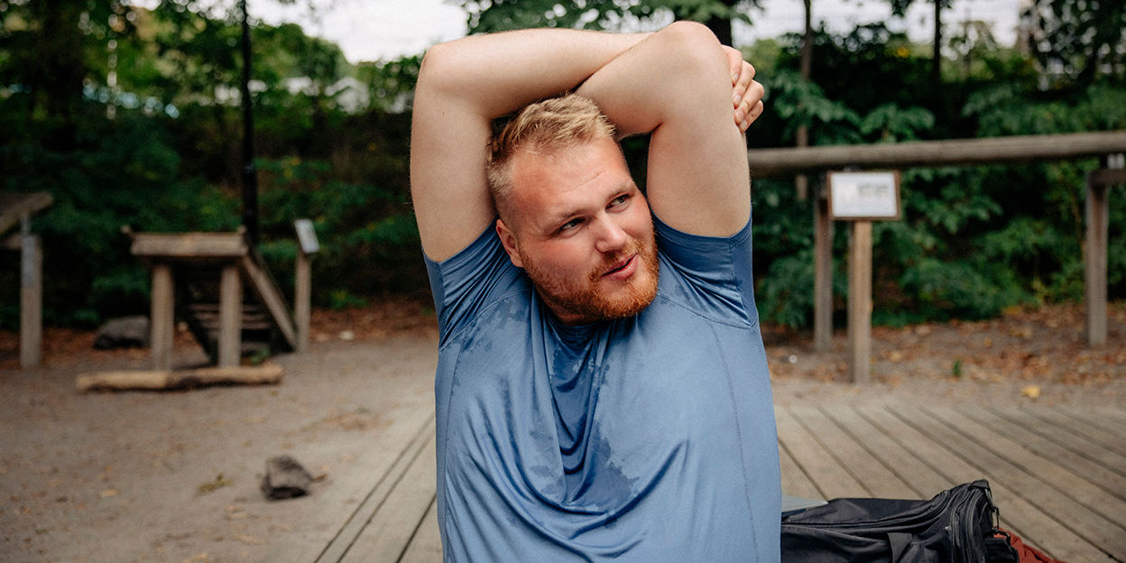 Kille sitter på ett utegym och håller upp armarna för att stretcha triceps. Han har en blå t-shirt som är lite svettig.