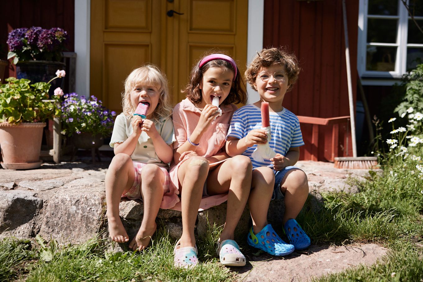 Tre barn sitter ute i solen på sommaren och äter glass i trädgården.