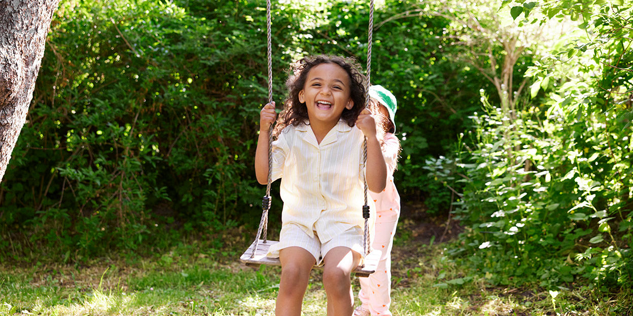 Två barn leker utomhus en solig dag . En sitter på gungan och den andra ger fart.